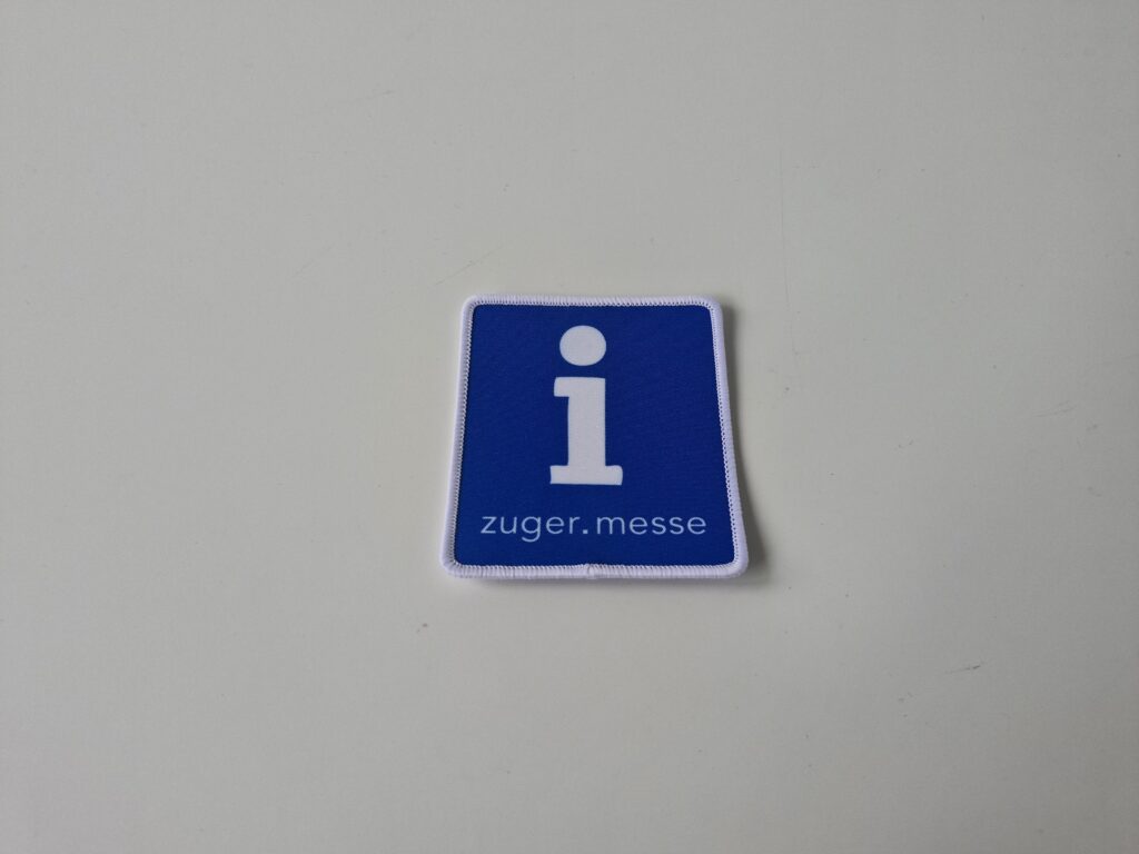 Bedruckter Aufnäher in Blau mit weissem Informationssymbol und Schriftzug „zuger messe“, mit weissem Kettelrand, auf neutralem Hintergrund fotografiert.