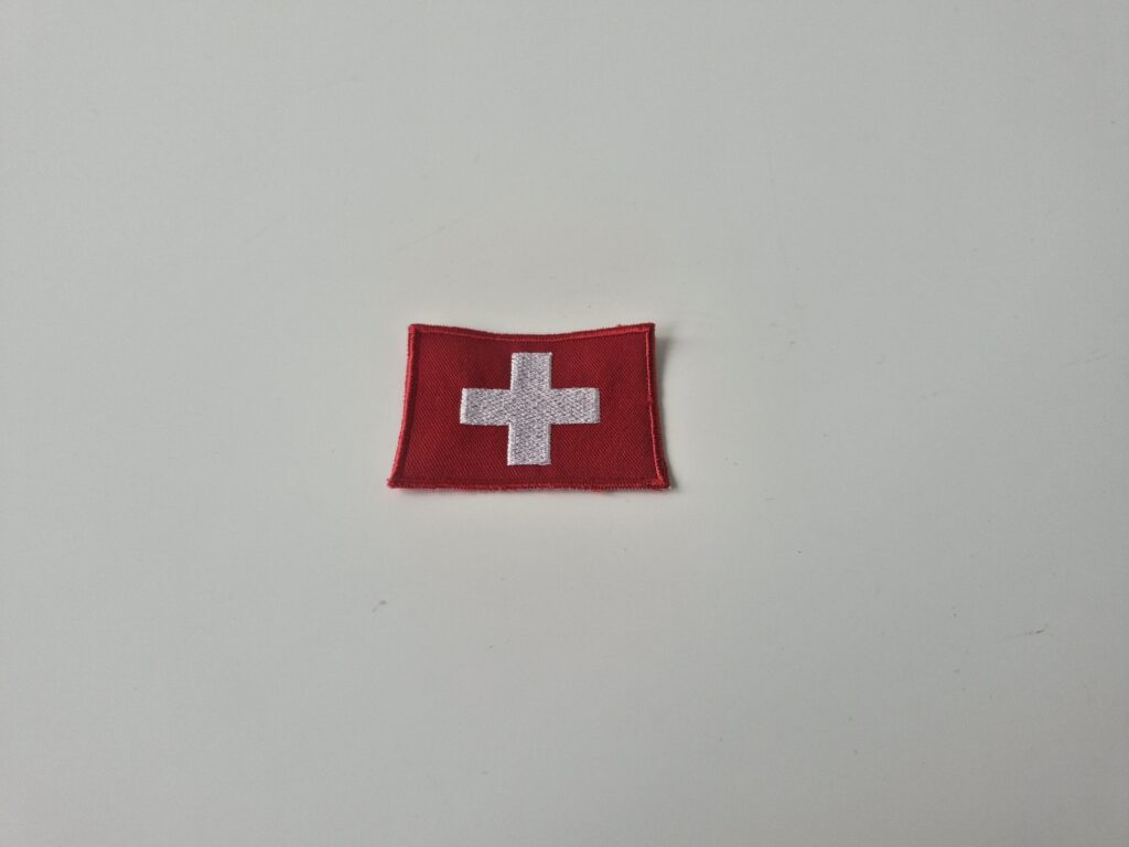 Gestickter Aufnäher in Rot mit weissem Kreuz (Schweizer Kreuz), fein bestickt und mit rotem Kettelrand, auf neutralem Hintergrund fotografiert.