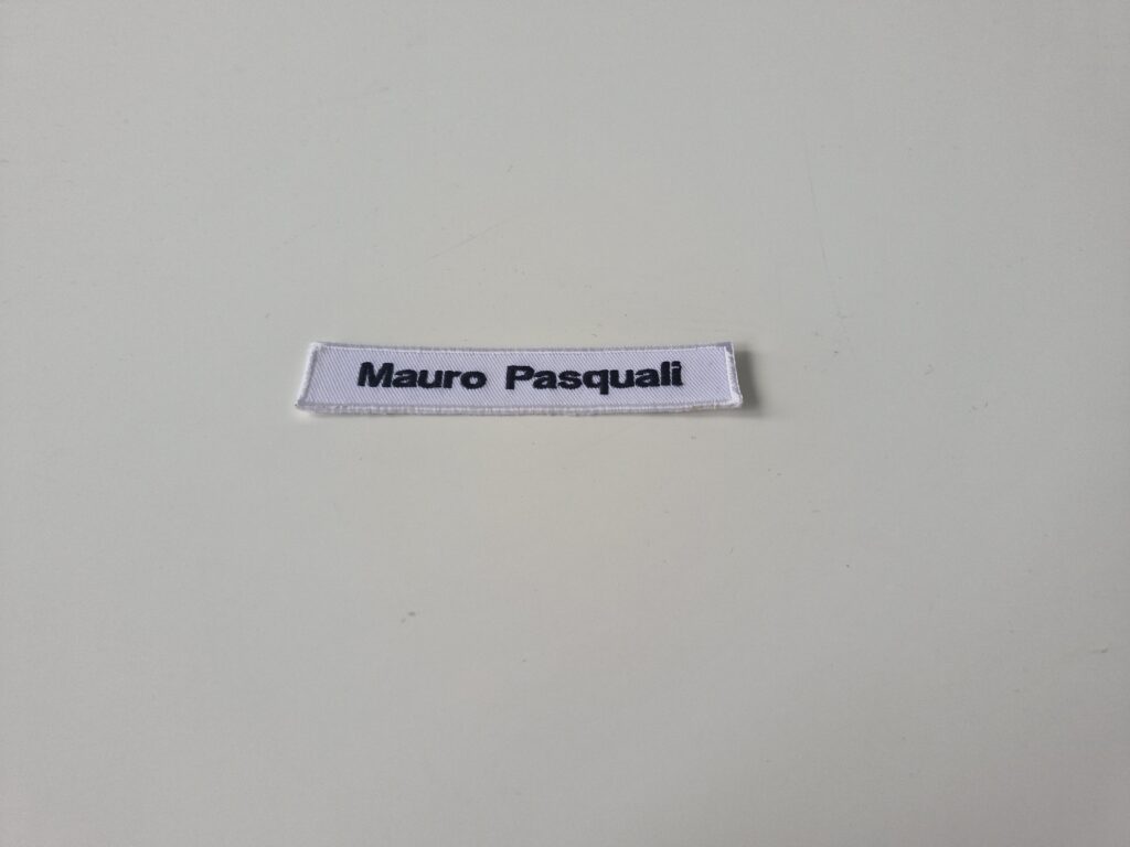 Weisser Namensschild-Aufnäher mit schwarzem Schriftzug „Mauro Pasquali“, rechteckig und sauber eingefasst, auf neutralem Hintergrund fotografiert.
